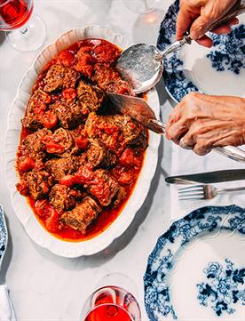 Image of a platter of beef braciole on white platter on table surrounded by blue and white plates and nonna hands serving